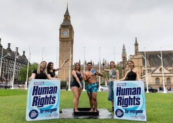 LUSH and EachOther activists stage a shower protest in parliament square. Two activists stand having a shower in their swimming costume and trunks, while others hold a shower head over them and big box mock ups of LUSH's 'human rights' product that reads: "human rights for everyone"
