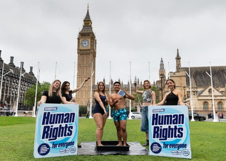 LUSH and EachOther activists stage a shower protest in parliament square. Two activists stand having a shower in their swimming costume and trunks, while others hold a shower head over them and big box mock ups of LUSH's 'human rights' product that reads: "human rights for everyone"