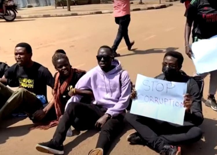 Ugandan anti-corruption protesters sit on the ground with placards.