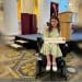Anna Landre in her wheelchair pointing at a stage which only has steps leading up to it. She is holding her Freedom of the City of London award, in an old fashioned room with large white pillars and patterned carpet. Anna is wearing a green patterned dress and white shoes and she looks sad