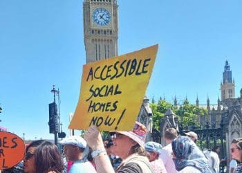 Paula Peters holding a yellow placard that reads in black writing "accessible social homes now" with Big Ben in the background. It is a sunny day at a protest over accessible housing