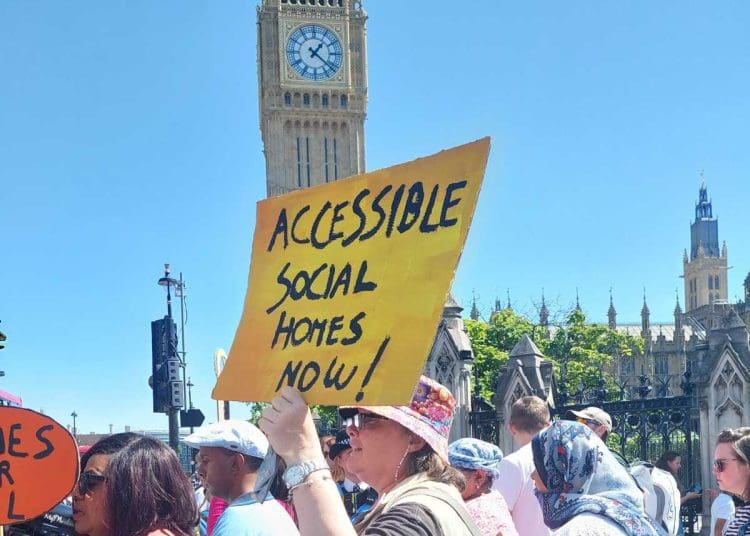 Paula Peters holding a yellow placard that reads in black writing "accessible social homes now" with Big Ben in the background. It is a sunny day at a protest over accessible housing