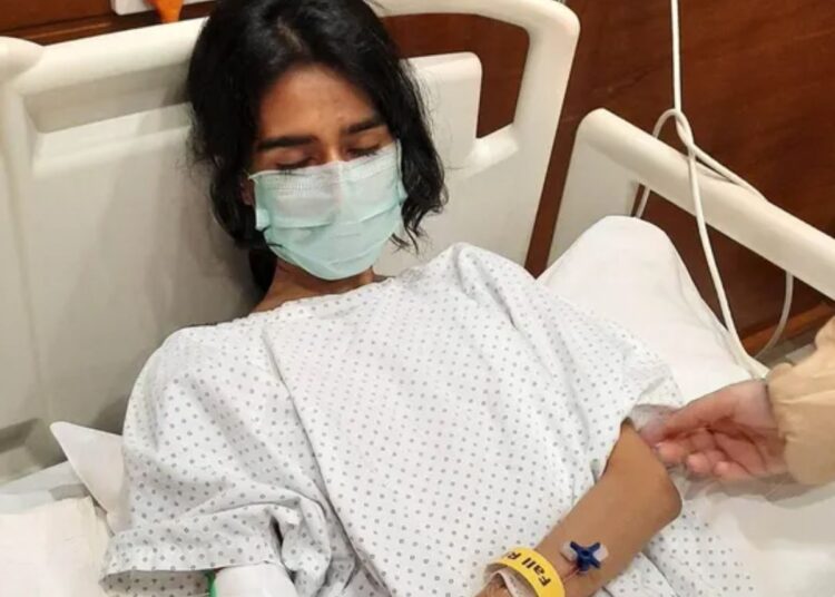 Severe ME/CFS patient Nevra in a hospital bed with her eyes closed, wearing a face-mask, and IV cannula attached to her wrist Pakistan