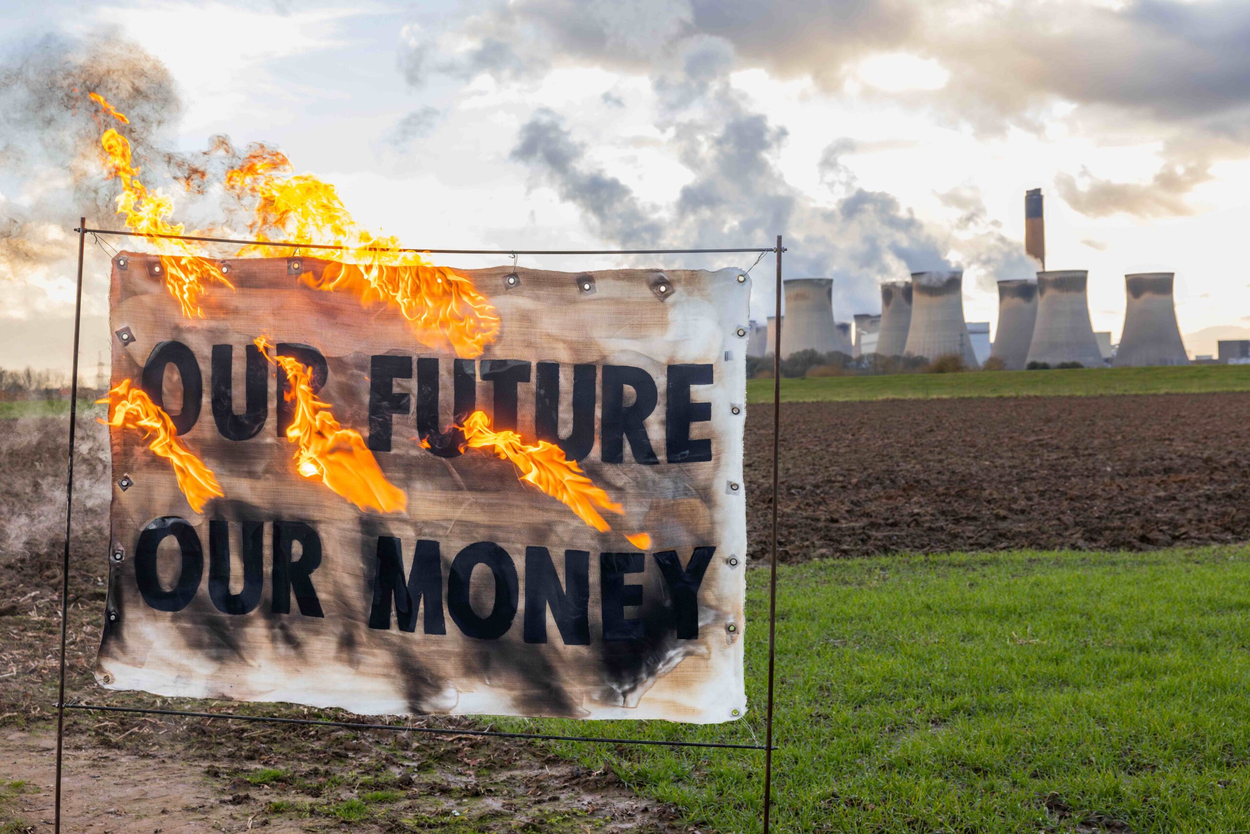 Banner on fire in front of Drax power station that reads: "Our future - our money", with smoke rising in the background from the smokestacks.