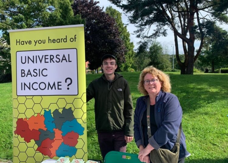 Louis Strappazzon and Alison Hawdale from the UBI Lab Network stand behind a table with campaign materials. An upright banner beside them reads: "Have you heard of a Universal Basic Income?"