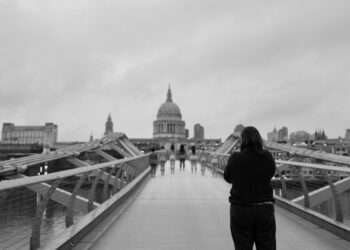 Grayscale image of St Paul's Cathedral in the background. Seven women/non-binary people stand nude with their backs to a photographer, in a line across Millennium Bridge photoshoot