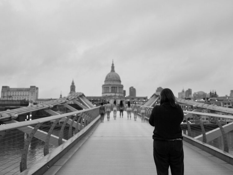 Grayscale image of St Paul's Cathedral in the background. Seven women/non-binary people stand nude with their backs to a photographer, in a line across Millennium Bridge photoshoot