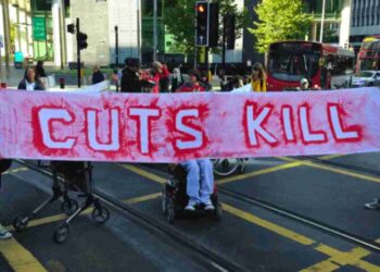 People holding a banner and blocking a road. The red writing on the banner reads "cuts kill". It was a protest over DWP cuts