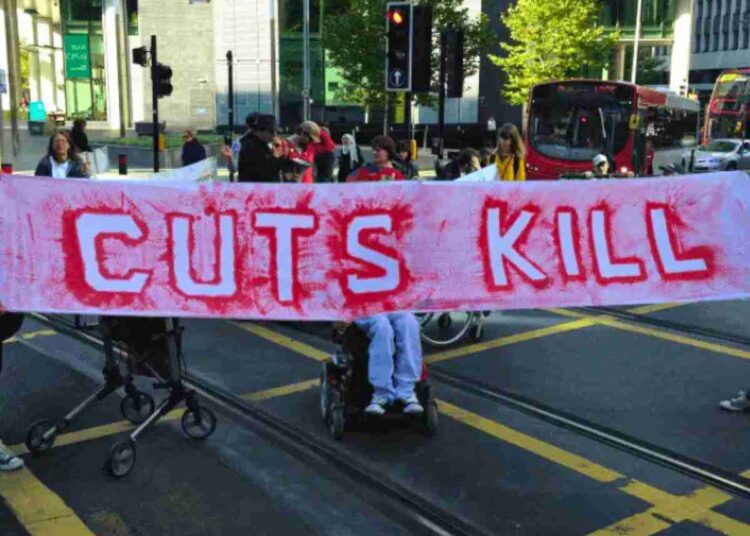 People holding a banner and blocking a road. The red writing on the banner reads "cuts kill". It was a protest over DWP cuts