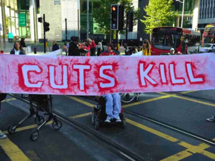 People holding a banner and blocking a road. The red writing on the banner reads "cuts kill". It was a protest over DWP cuts