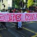 People holding a banner and blocking a road. The red writing on the banner reads "cuts kill". It was a protest over DWP cuts
