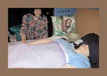 Karen Gordon, a severe ME patient, laying in bed with her eyes and ears covered as her mother looks on