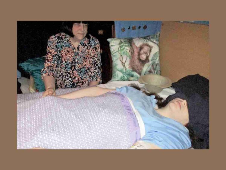Karen Gordon, a severe ME patient, laying in bed with her eyes and ears covered as her mother looks on