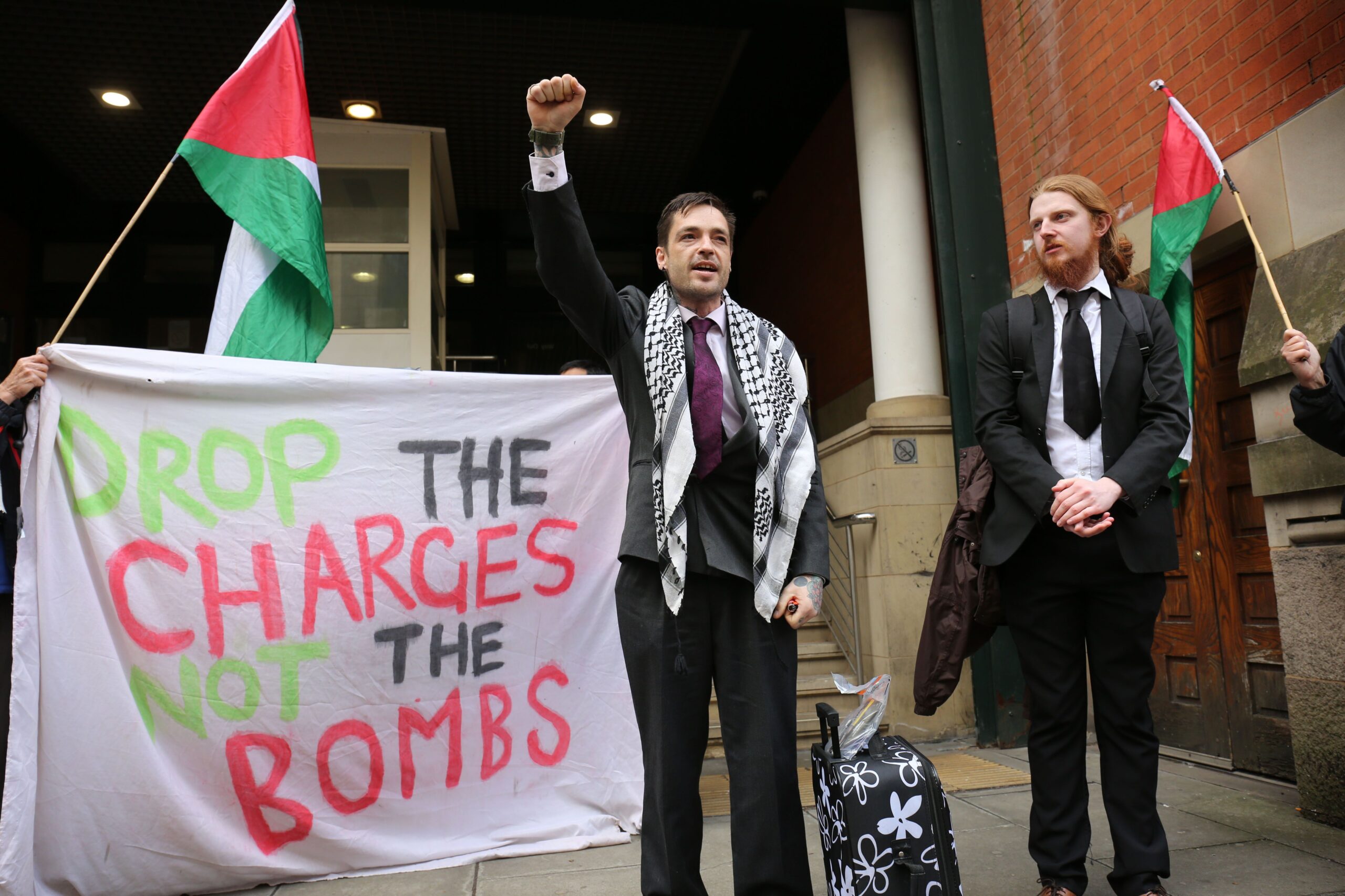 Palestine Action Ferranti 2 activist stands with a raised fist, wearing a suit and keffiyeh, next to Ferranti 2 activist Adam, while protesters with Palestine flags and a banner reading: "Drop the charges, not the bombs" behind them. 