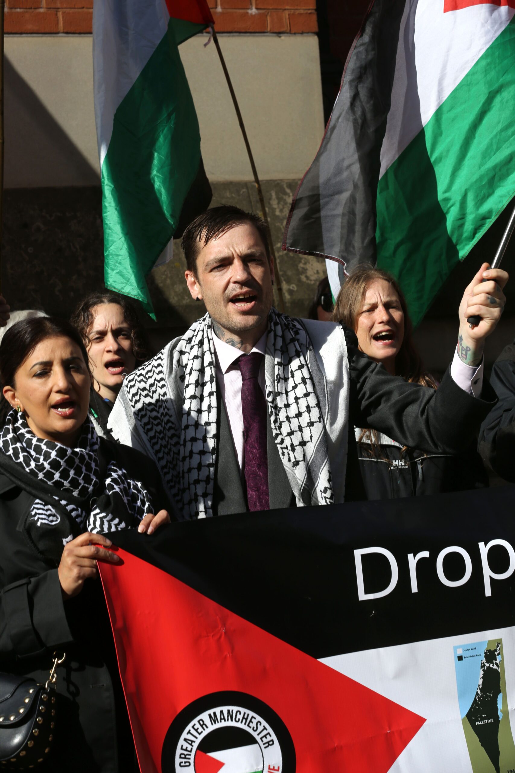 Palestine Action Ferranti 2 activist Drew stands amid a crowd of supporters holding banners and flags outside court. 