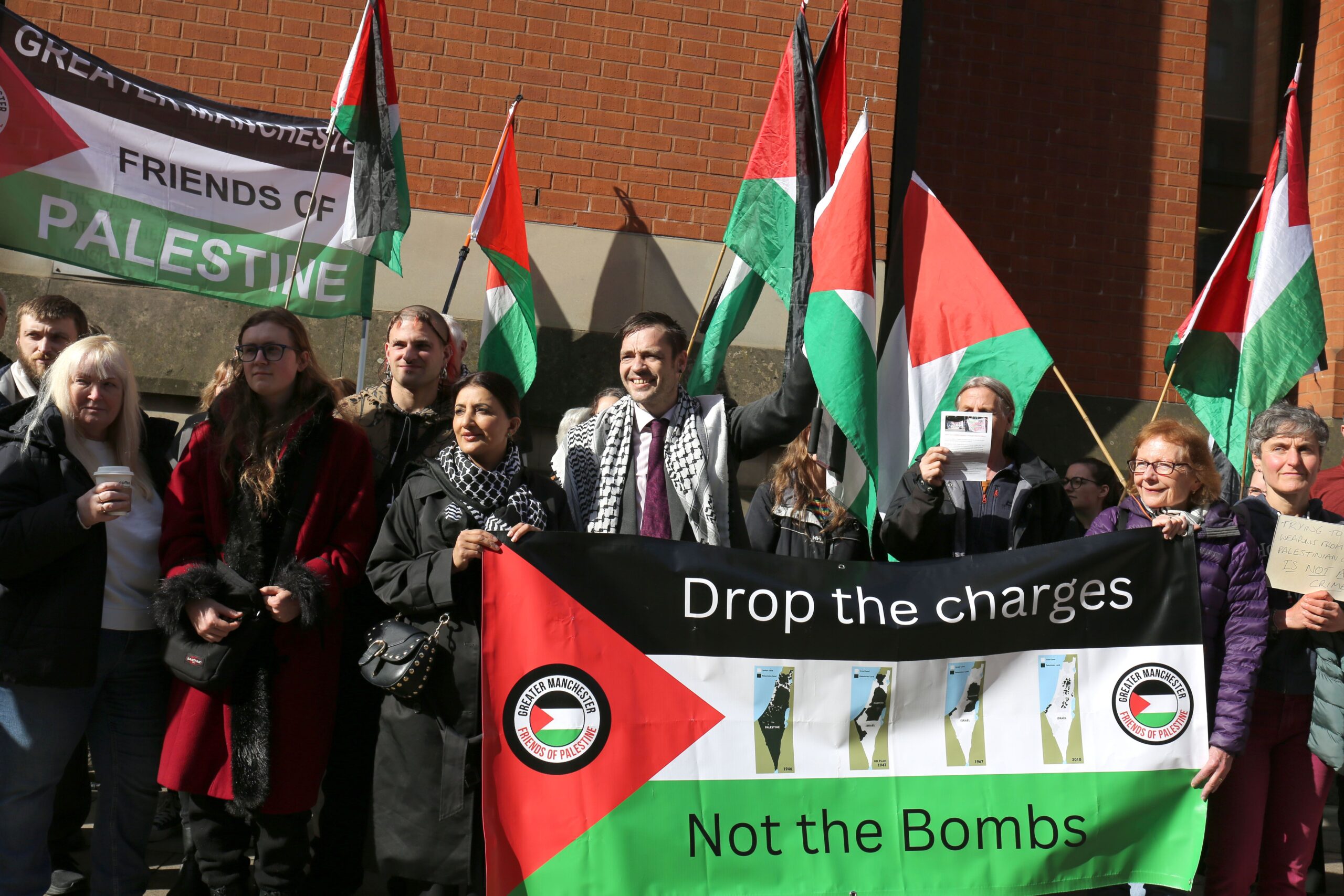 Large group of protesters holding Palestine flags, including one which reads: "Drop the charges, not the bombs" with four illustrations of Palestine showing increasing Israeli annexation. Defendant Drew stands wearing a suit and keffiyeh in the centre. 