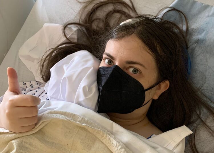 Emily in a hospital bed after surgery making a thumbs up, wearing a black mask, and with a dressing on her head. Trump Medicare