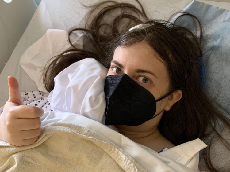 Emily in a hospital bed after surgery making a thumbs up, wearing a black mask, and with a dressing on her head. Trump Medicare