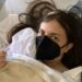 Emily in a hospital bed after surgery making a thumbs up, wearing a black mask, and with a dressing on her head. Trump Medicare