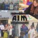 Autistic Inclusive Meets (AIM) logo over four images. Top Left: campaigners stand behind a table protesting ABA therapy at Ambitious About Autism's fundraising event in London. Bottom left: Four members of AIM's staff stand outside Number 10 with a guard accepting a petition. Top right: Young white girl with long hair and headphones on colours in the rainbow autism infinity symbol. Bottom right: Three black kids play together with a sensory table with a their mum watching over.