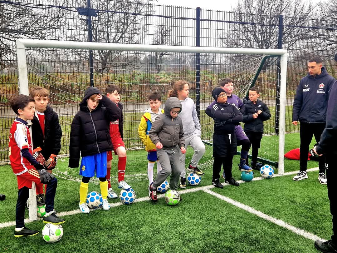 Children in a line across a goal, with a football each at their feet. 