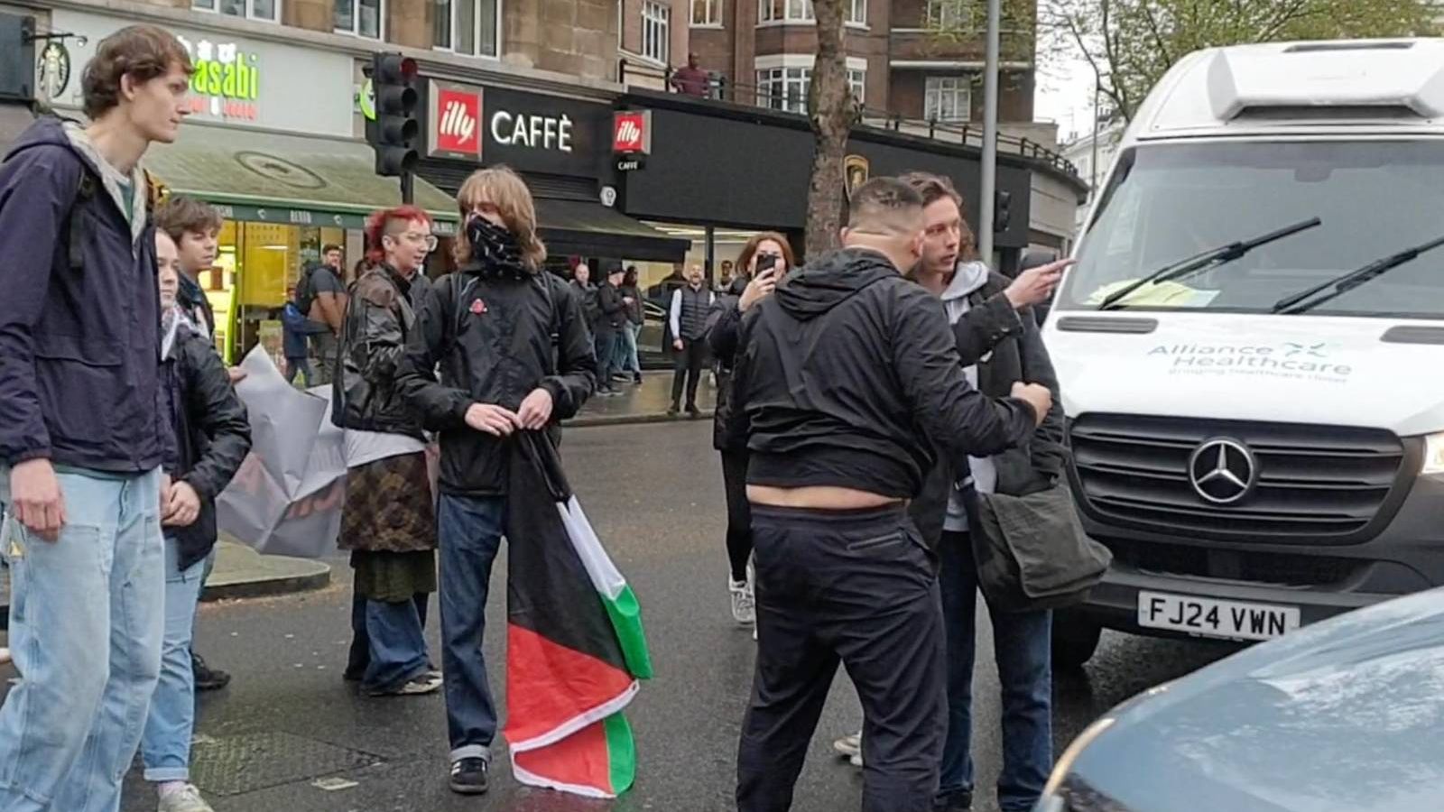 A white male passerby confronts Youth Demand protesters, who are stood blocking traffic. 