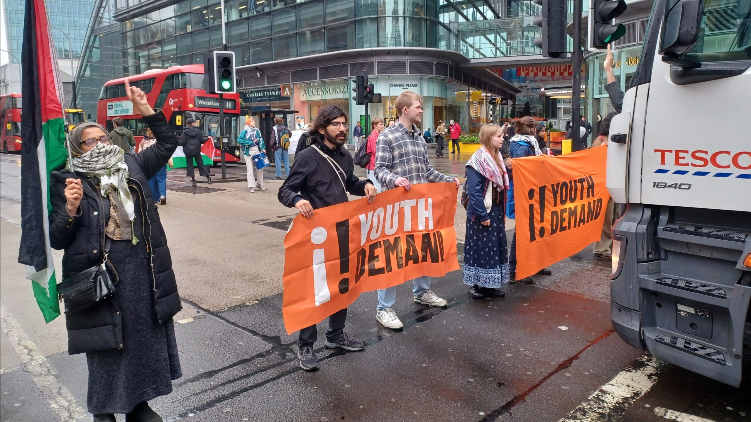 protesters, some wearing keffiyehs, stand in a line with orange banners blocking oncoming traffic. One holds up a Palestine flag.