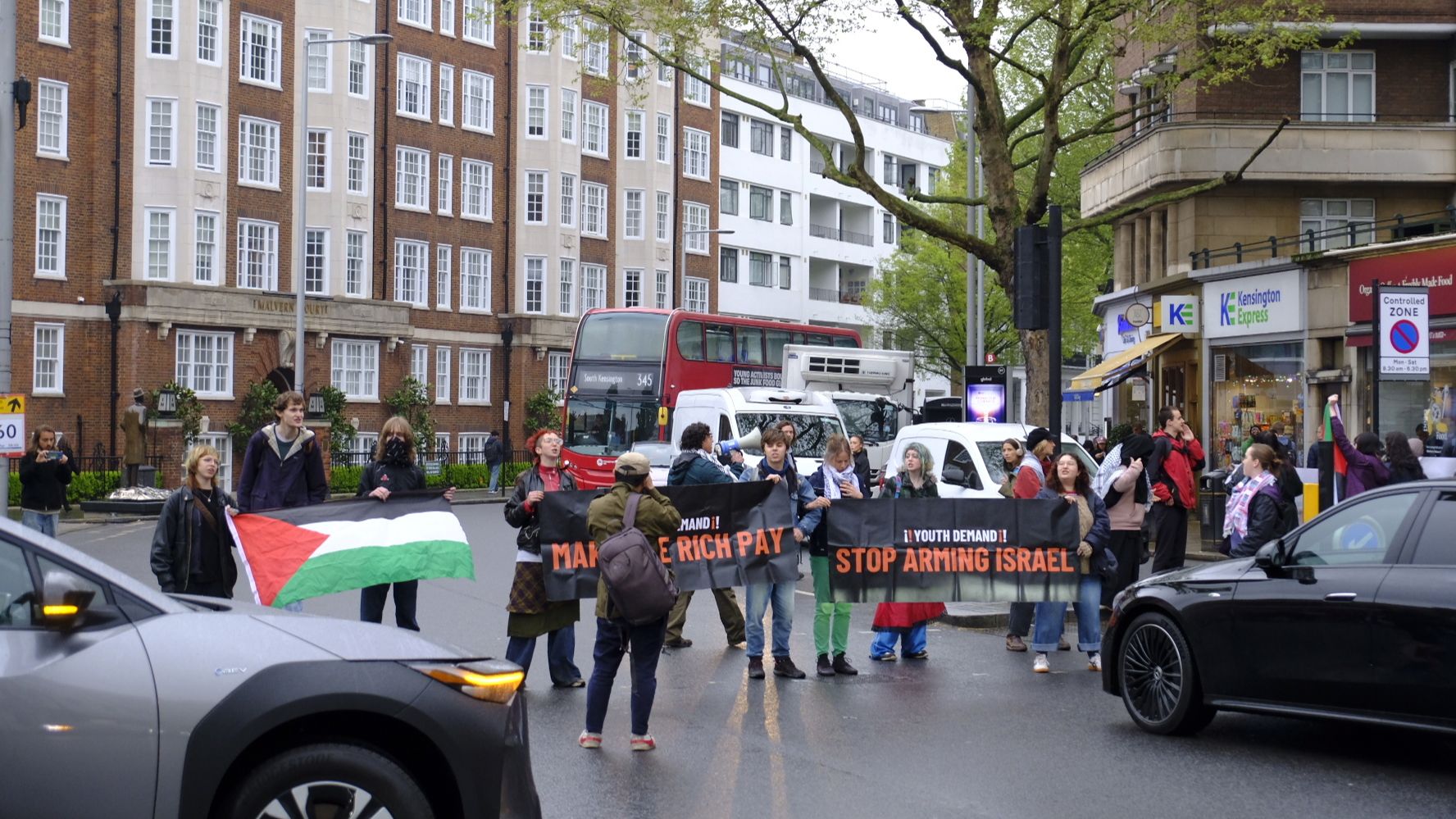 Youth Demand activists stand in a line in front of cars, holding banners with the group's logo and "Stop arming Israel" and "Make the rich pay".