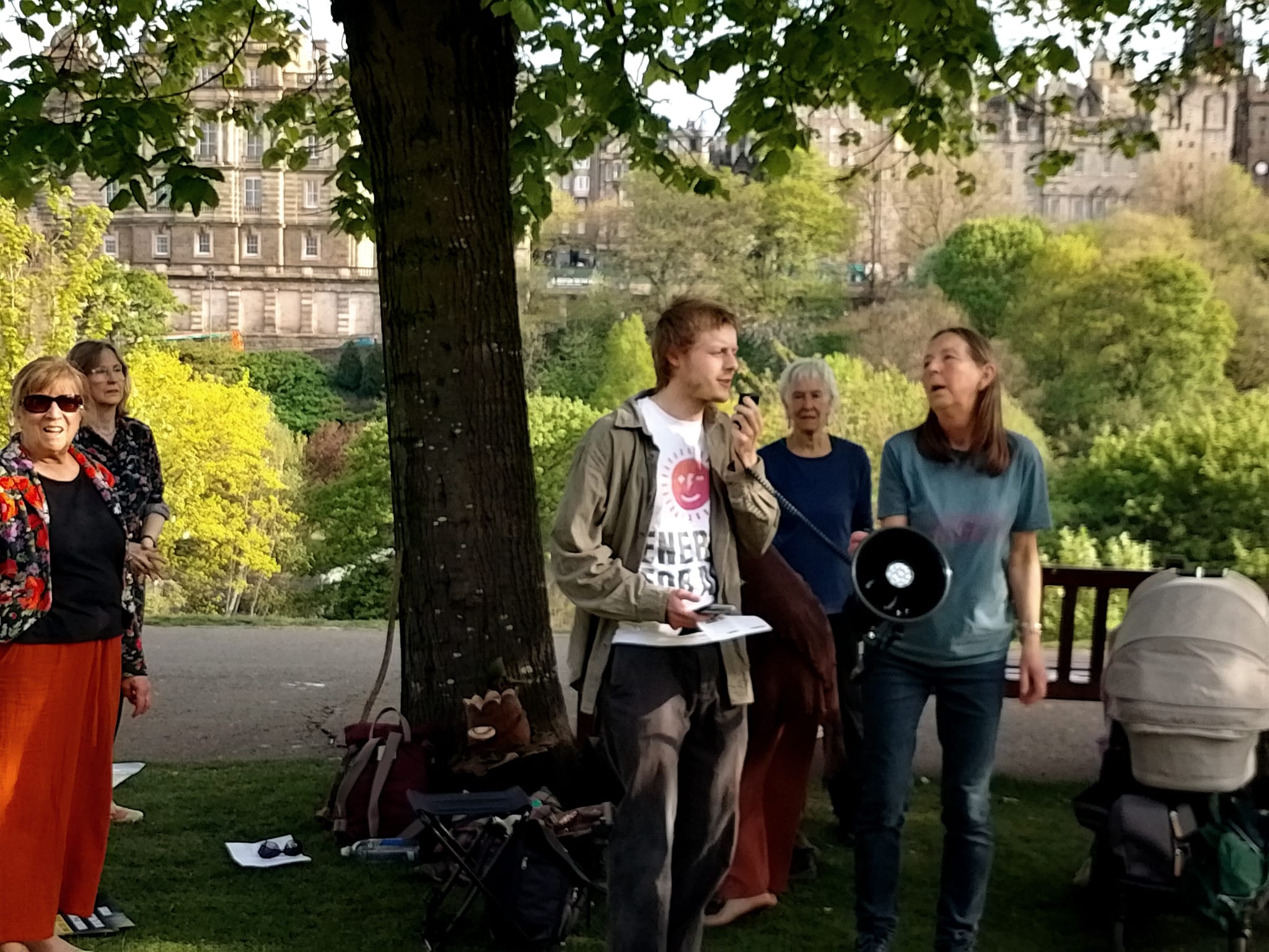 Drax AGM demo Edinburgh. Stuart Bretherton from Fuel Poverty Action giving a speech under a tree, with other campaigners looking on. 