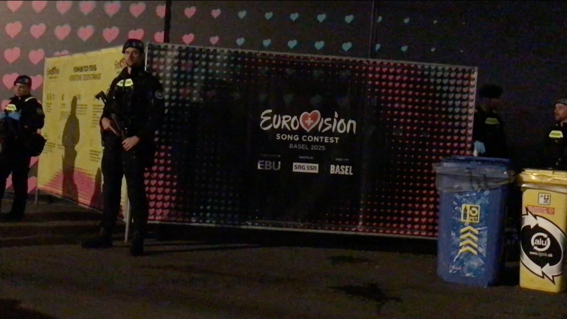 Youth Demand supporters behind a Eurovision screen, guarded by two armed police. 