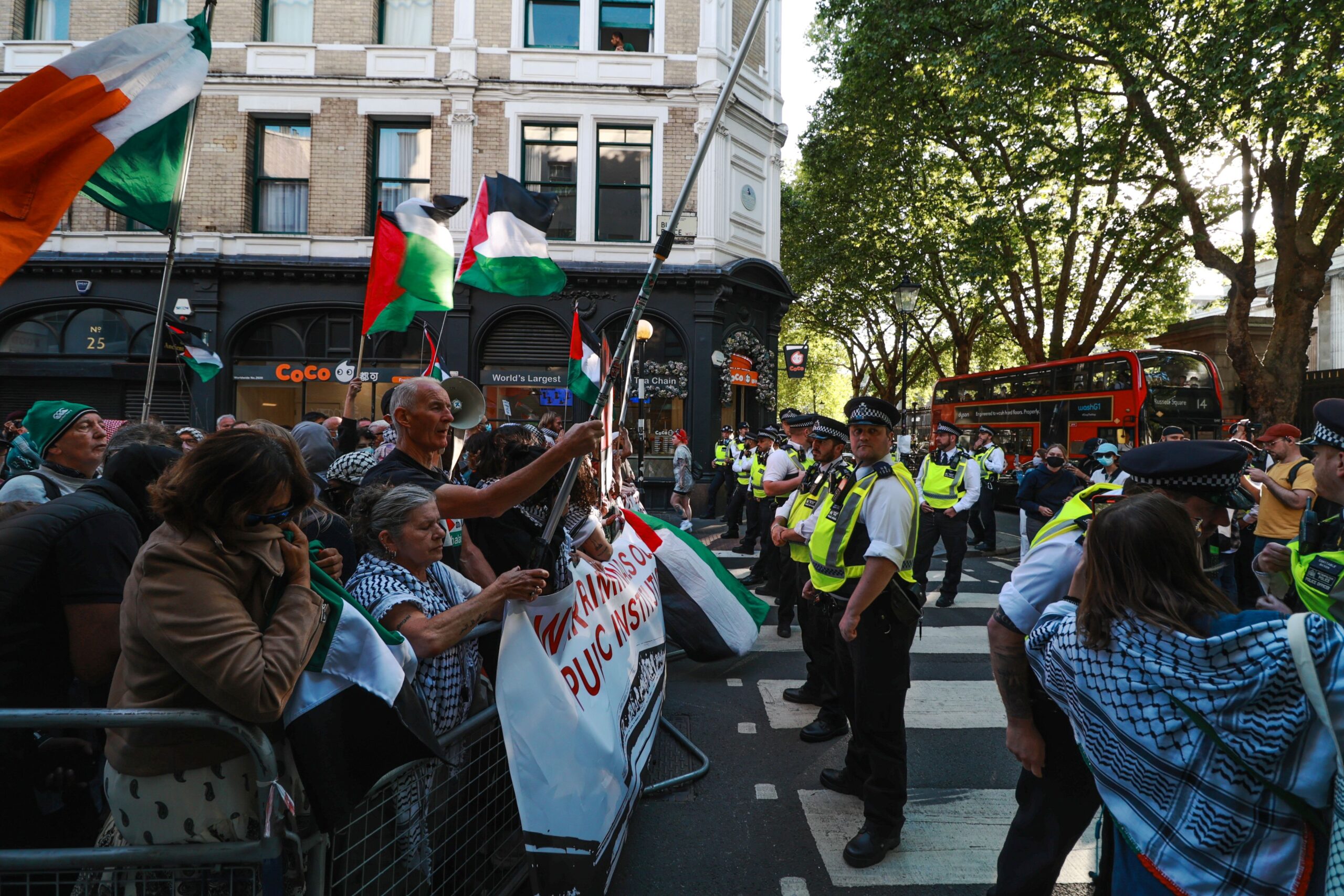 A large number of police form a line in front of Palestine protesters holding flags and banners behind barricade fences. 