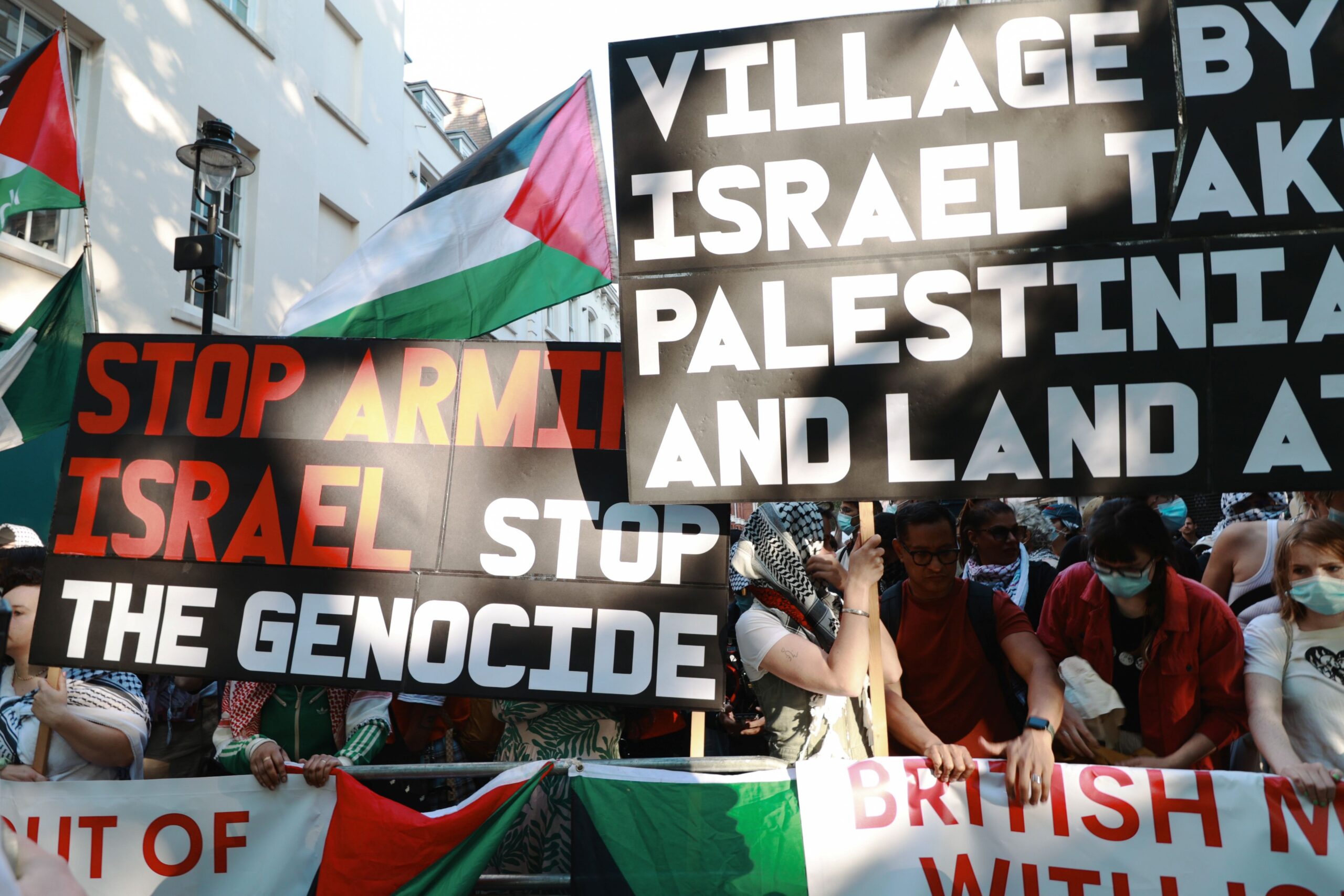 Protesters in a crowd raise banners and placards reading "Stop arming Israel, stop the genocide" and "Village by village, Israel takes Palestinian homes and land at gunpoint". 