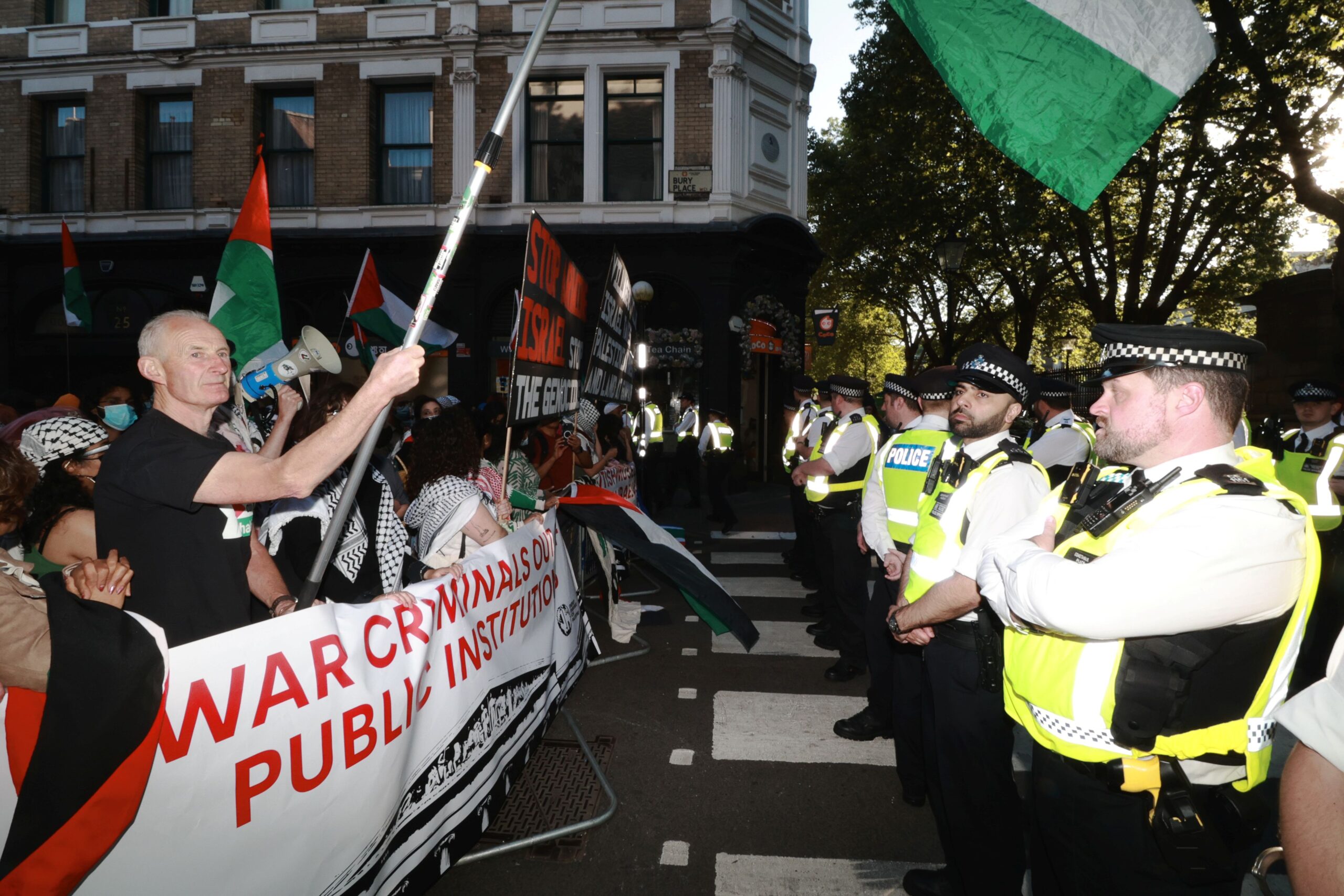 A large number of police form a line in front of Palestine protesters holding flags and banners. 
