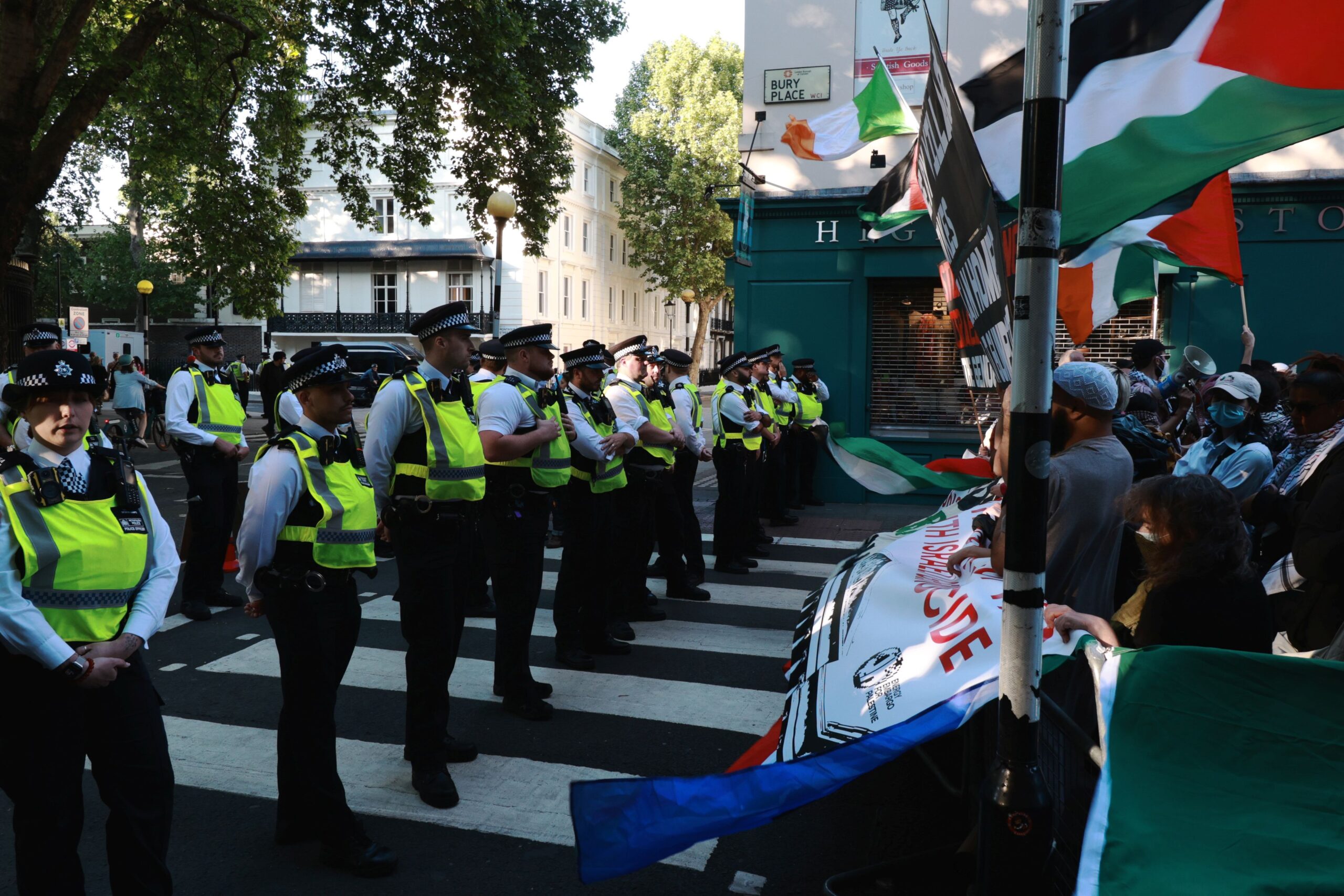 A large number of police form a line in front of Palestine protesters holding flags and banners. 