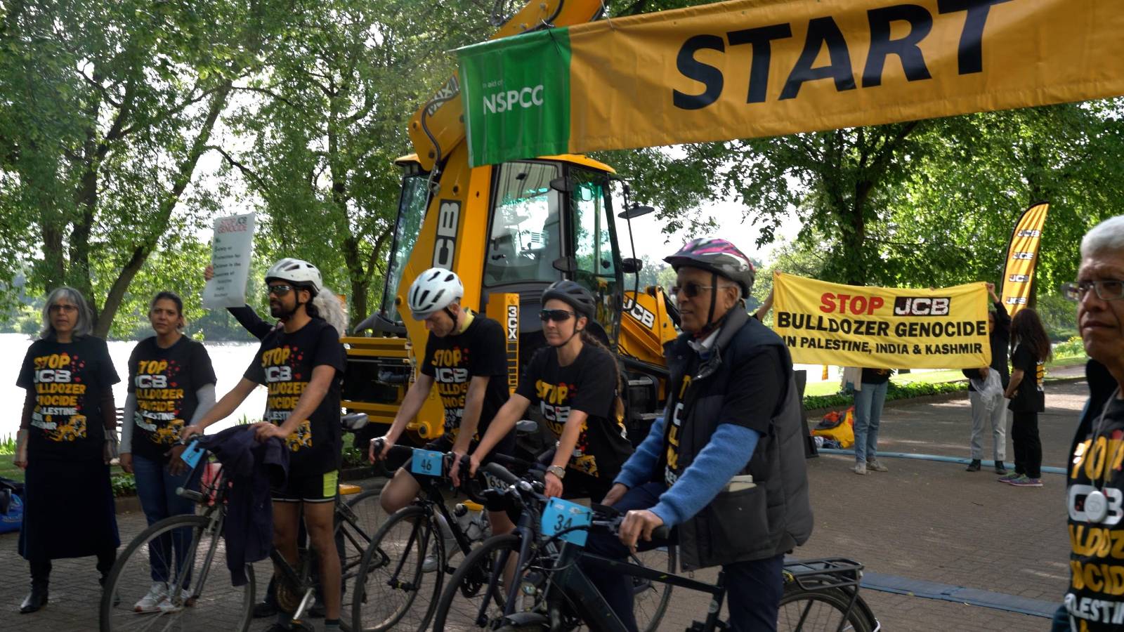 Cyclists and supporters wearing "Stop JCB's Bulldozer genocide In Palestine, India and Kashmir" T-shirts stand at the starting line together, with a group holding a banner reading: "Stop JCB bulldozer genocide in Palestine, India & Kashmir". 