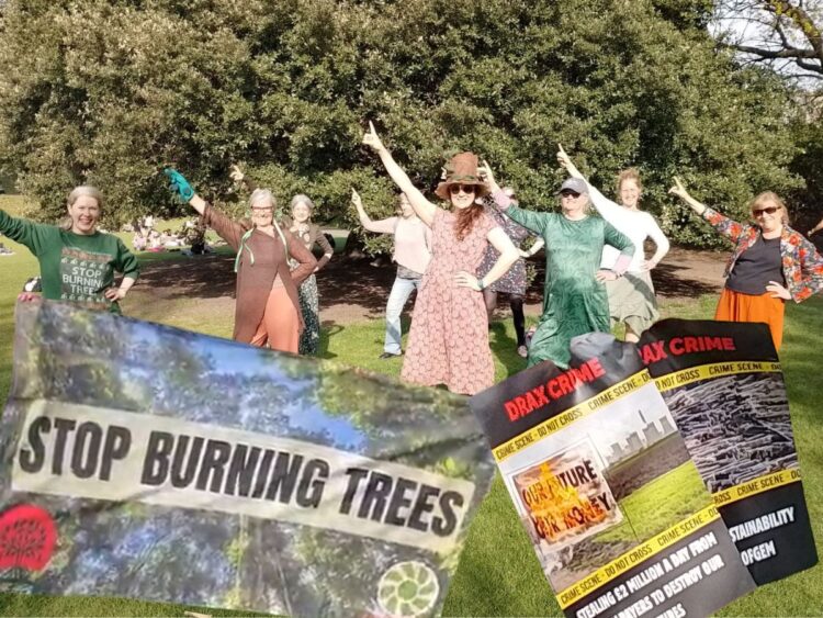 Nine women strike a pose while dancing on the grass, with trees behind, in Princes Street Gardens, Edinburgh. Stop burning trees banner and Drax crime posters superimposed over the top Drax AGM