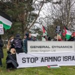Protesters with Palestine flags and banners reading "Stop arming Israel" stand next to General Dynamics' sign.