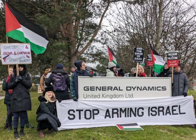 Protesters with Palestine flags and banners reading "Stop arming Israel" stand next to General Dynamics' sign.