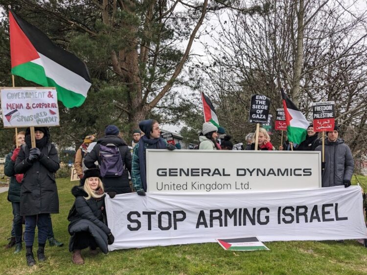 Protesters with Palestine flags and banners reading "Stop arming Israel" stand next to General Dynamics' sign.