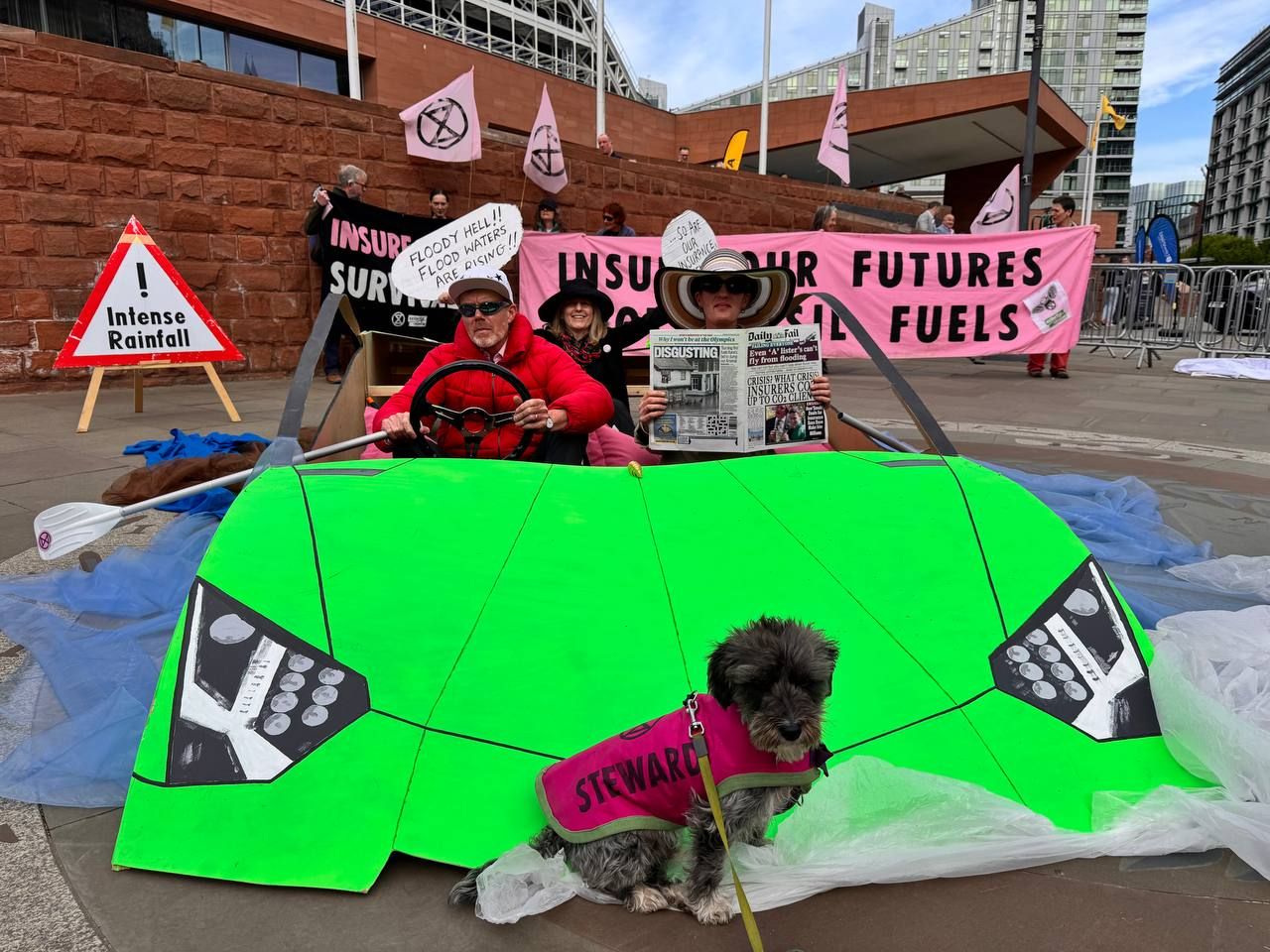 Vivid green model of a Lamborghini sports car, with three activists sat inside. One drives, another holds a 'Daily Fail' newspaper, and the last is holding a speech bubble placard reading: "Floody hell!! Flood waters are rising!!" Behind, activist hold a banner reading "insure our futures, not fossil fuels" and "Insure survival". A hazard sign with "Intense Rainfall" has been placed behind. A dog wearing a coat reading "steward" sits on the bonnet. Extinction Rebellion