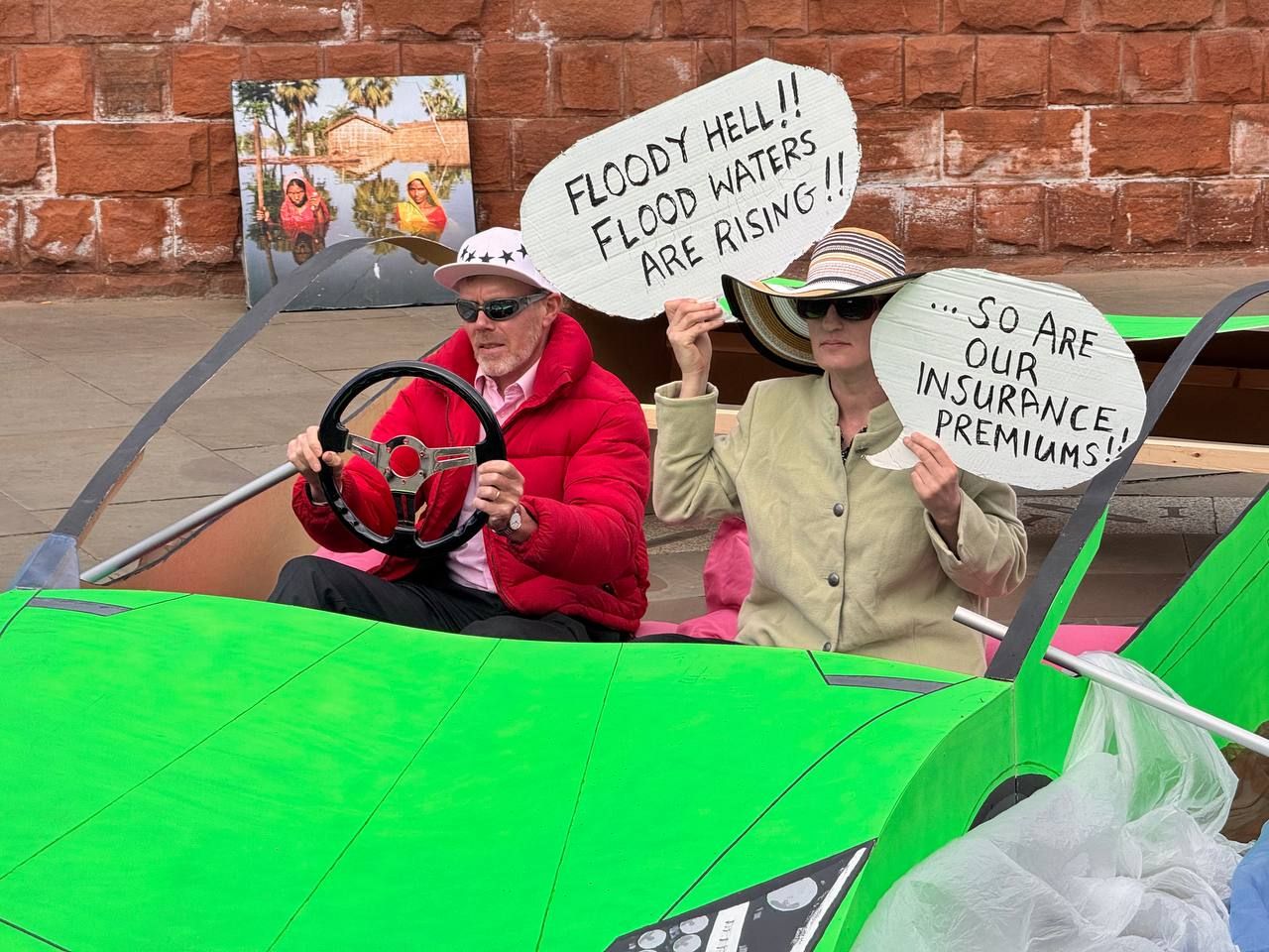 Vivid green Lamborghini with two activists inside. One drives while rowing, and the other holds speech bubble placards reading "Floody hell!! Flood waters are rising!!" and "So are our insurance premiums" Extinction Rebellion