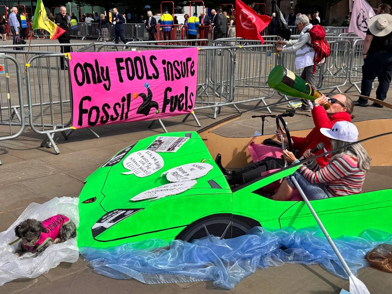 Vivid green Lamborghini with two activists inside rowing through flood waters. A bright pink banner behind reads: "Only fools insure fossil fuels"