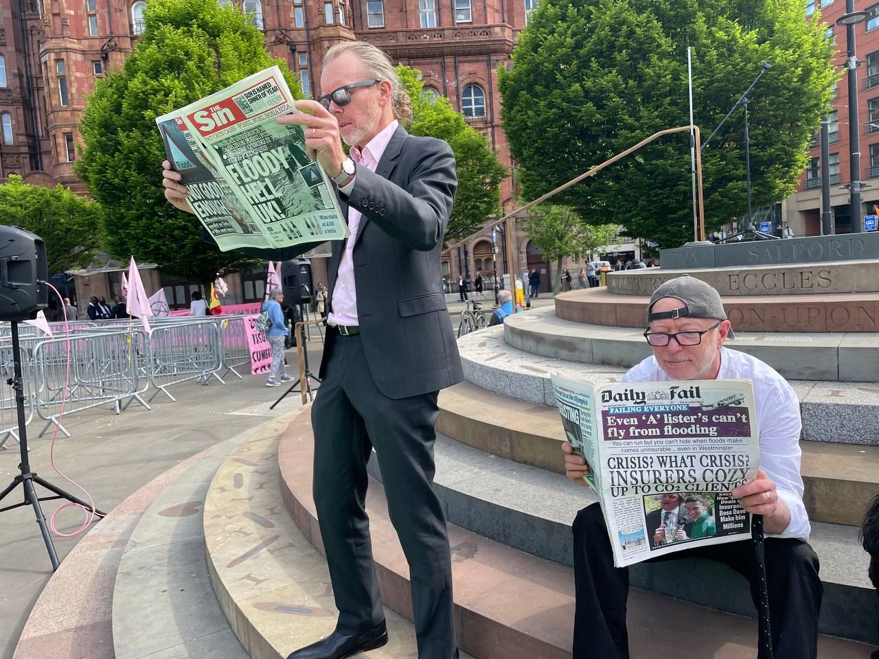 Two men hold and read mock newspapers The Sin and the Daily Fail. Headlines read: "Floody Hell UK!" and "Crisis? What crisis? Insurers cozy up to Co2 clients" Other text on them reads: "Even 'A' lister's can't fly from flooding" and "Not good enough" next to a flood sign. 