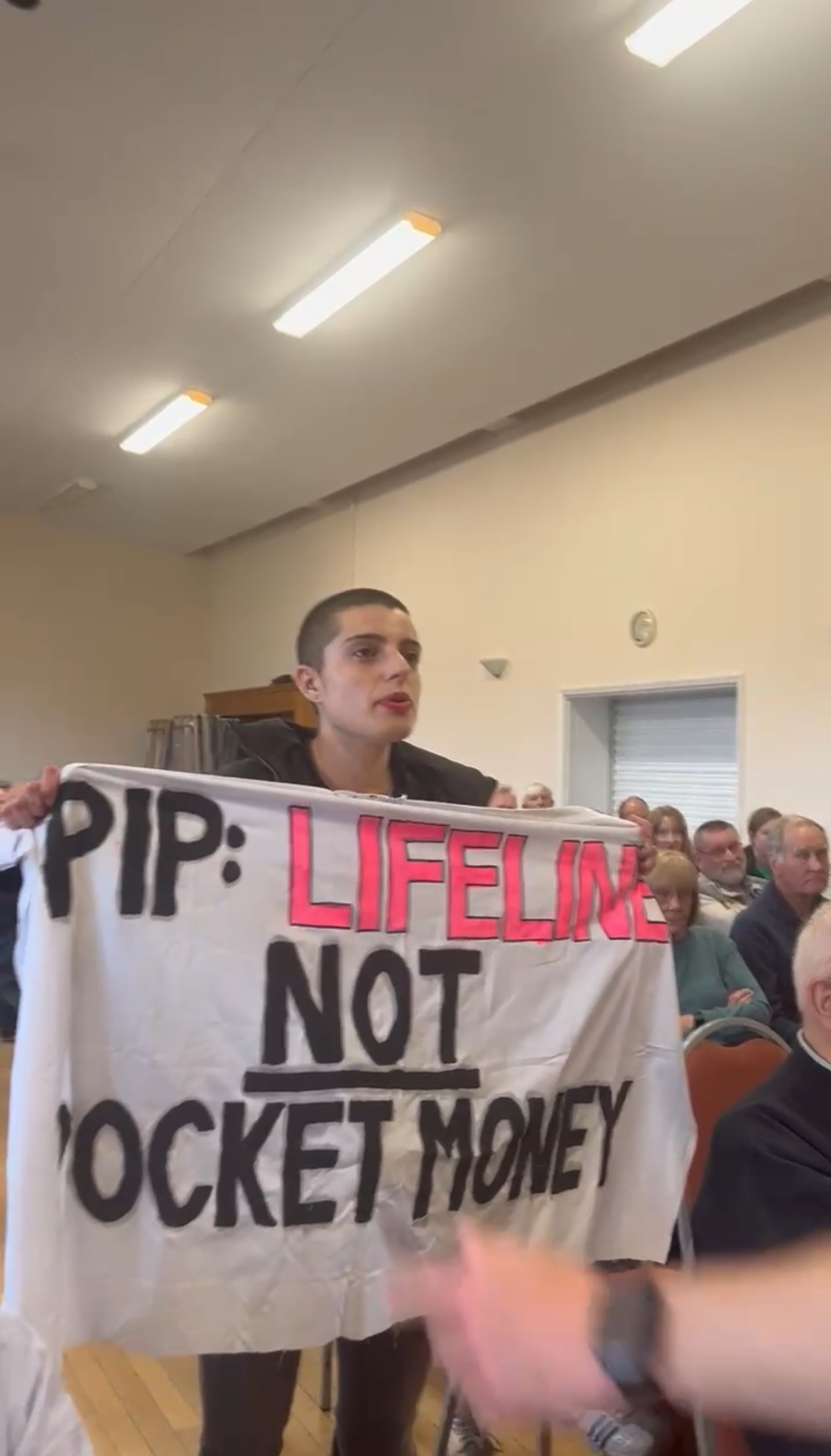 Gigi stands up in a packed town hall meeting with a banner reading: "PIP: Lifeline not pocket money".