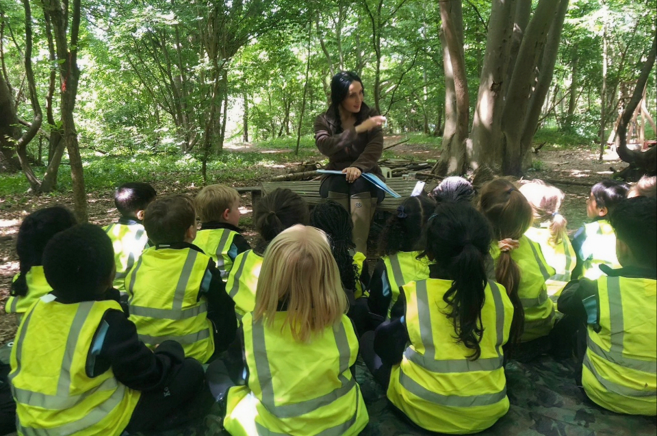 Carly Ahlen sits in the forest with a group of students - backs to the photo - in high vis vests, attentively listening to her talk. 