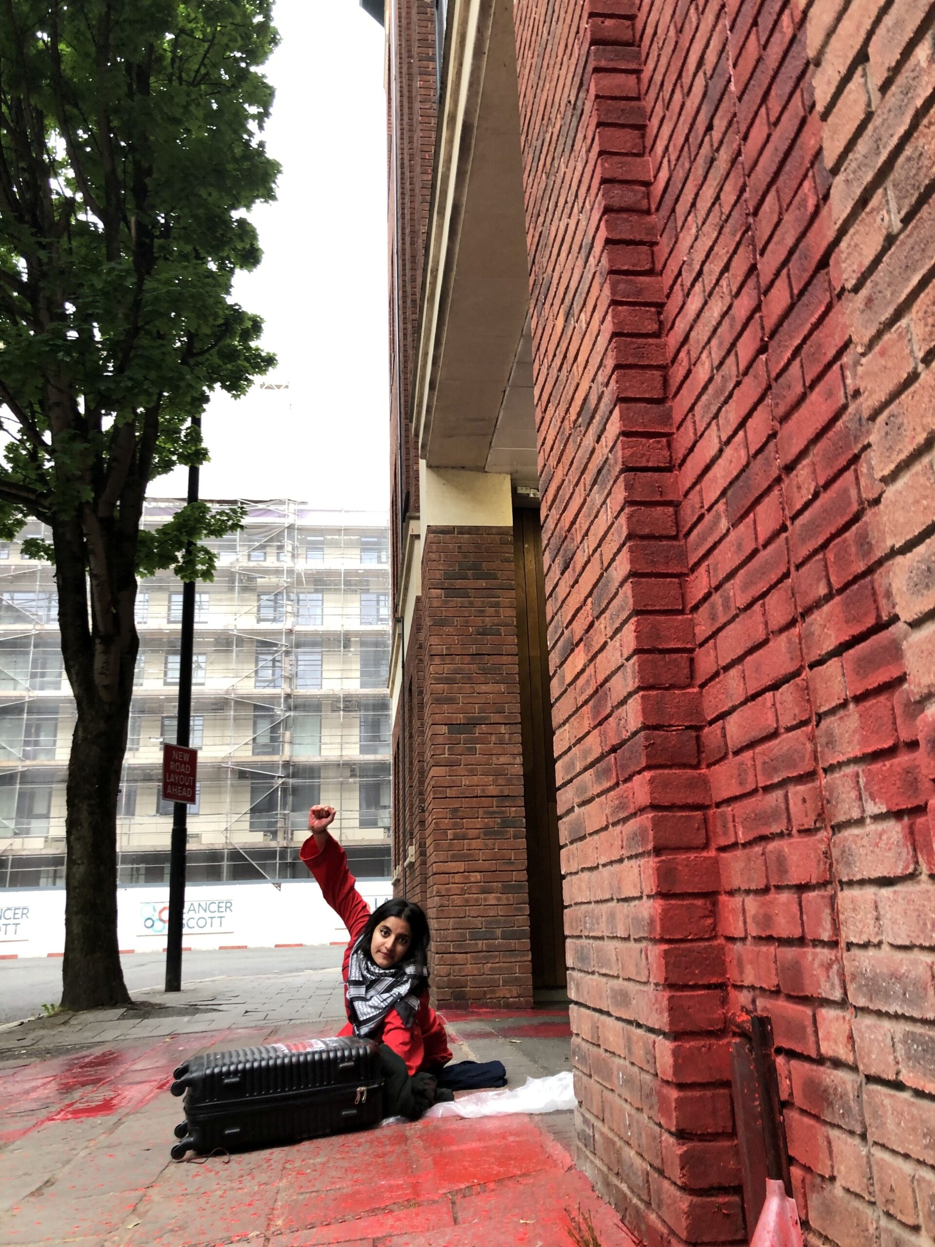 Palestine Action activist lies on the pavement attacked to a black and white striped suitcase, with a solidarity fist in the air, a keffiyeh round her neck, and red paint splatters across the pavement and wall surrounding her. 