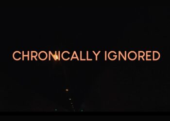 'Chronically Ignored' in orange text over a black background with a few small lights running down the ceiling of a pitch-black tunnel ME/CFS