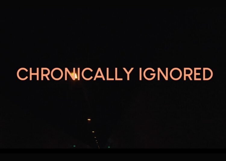 'Chronically Ignored' in orange text over a black background with a few small lights running down the ceiling of a pitch-black tunnel ME/CFS