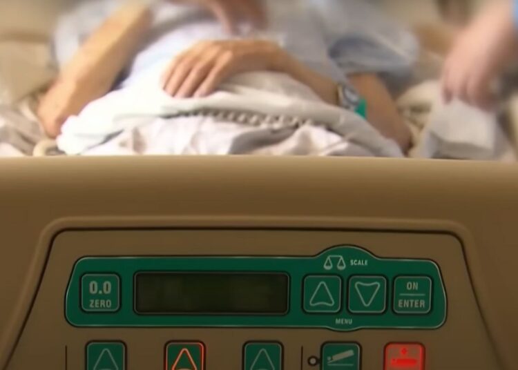 Face on view of a person in a hospital bed, from the foot of the bed. A medical practitioner stands next to them. Bill on assisted dying.
