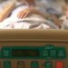 Face on view of a person in a hospital bed, from the foot of the bed. A medical practitioner stands next to them. Bill on assisted dying.